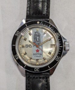 Orologio Vostok Komandirskie commemorativo 1945, quadrante argentato con statua della Vittoria, ghiera in bachelite nera, movimento meccanico manuale Vostok 2414, cassa in ottone cromato, cinturino sovietico originale.