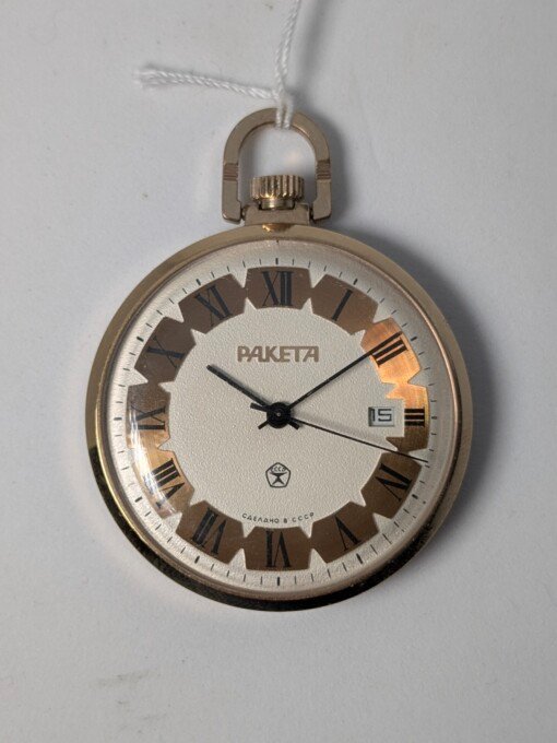 Orologio da tasca sovietico Raketa con cassa dorata, quadrante bianco, numeri romani neri alternati a indici lineari dorati, datario alle ore 3 e il marchio di qualità sovietico GOST.