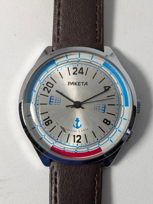Orologio da polso sovietico Raketa Marine 24 ore vintage degli anni '80 con quadrante argentato, indicazione delle 24 ore, lunetta blu e rossa con simboli marittimi e cinturino in pelle marrone.