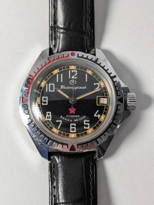 Orologio da polso militare sovietico Vostok Komandirskie Zakaz MO CCCP a carica manuale (calibro 2414) con quadrante nero, numeri arabi bianchi, lancette luminose, ghiera girevole e cinturino in pelle nera.